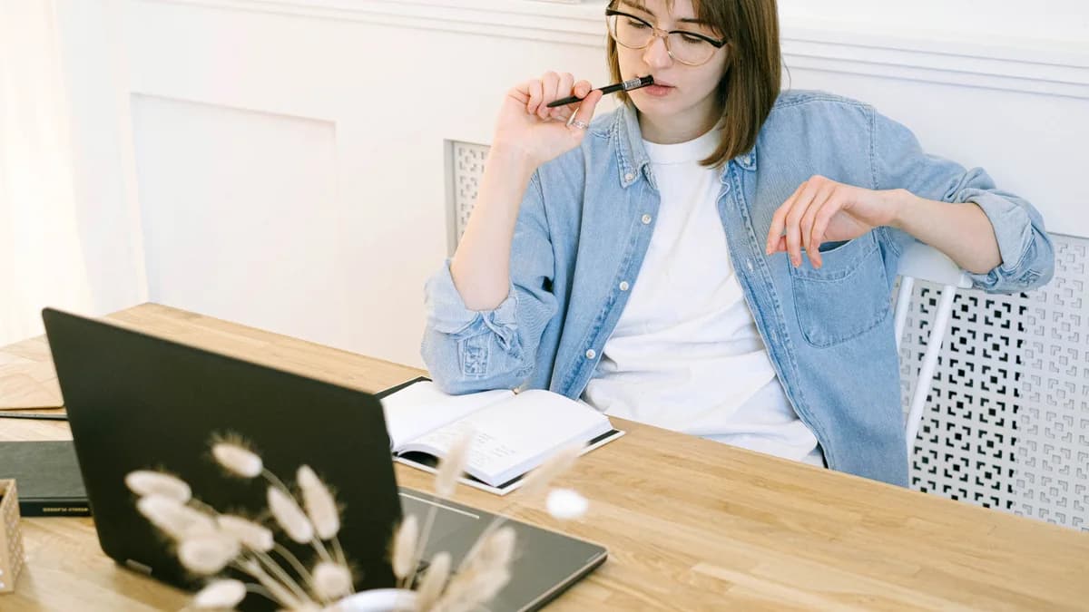 Frau mit Brille sitzt nachdenklich am Schreibtisch vor einem Laptop und einem Notizbuch