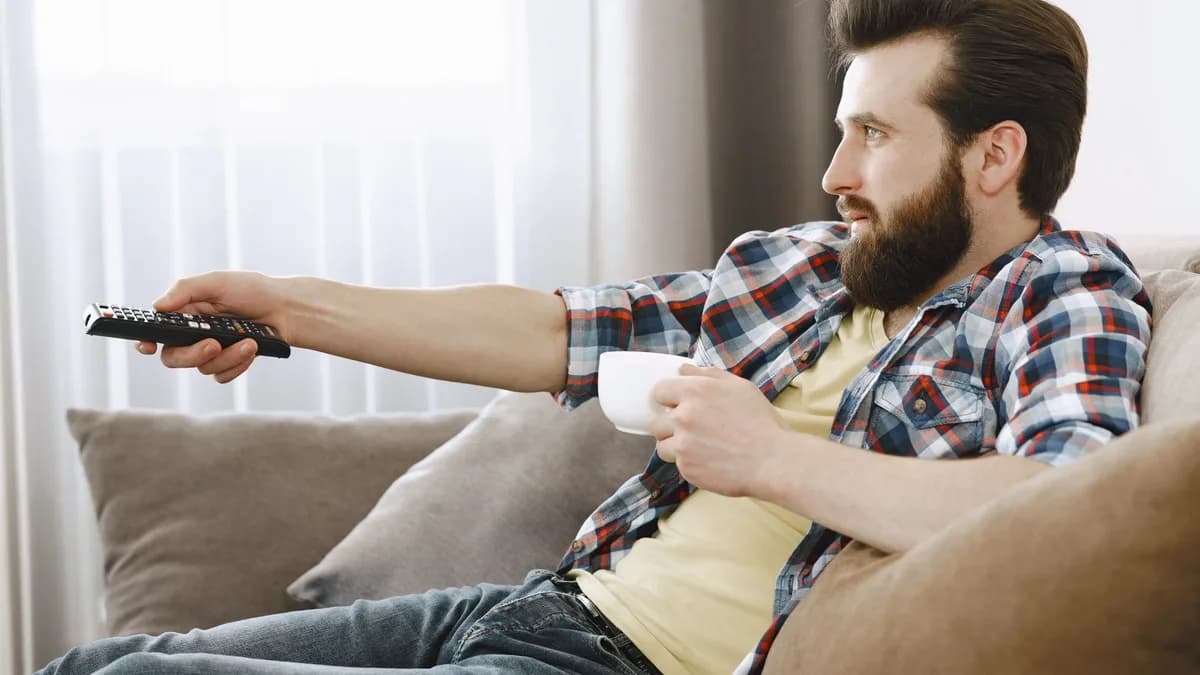 Mann entspannt auf dem Sofa mit Fernbedienung und Kaffeetasse in der Hand